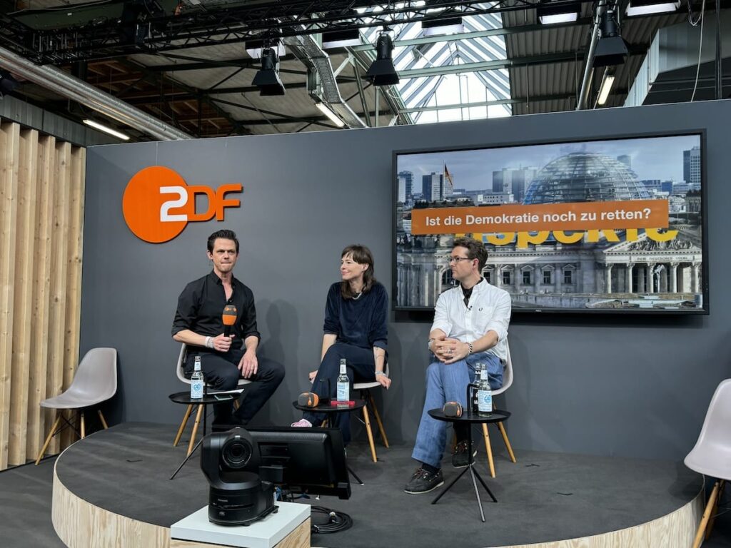 re:publica24-Panel "Ist die Demokratie noch zu retten?" im ZDF Meet Up Corner - Jo Schück, Hedwig Richter, Veith Selk (von links) sitzen auf Stühlen auf einer kleinen Bühne, links dahinter das ZDF-Logo, rechts ein großer Monitor. 